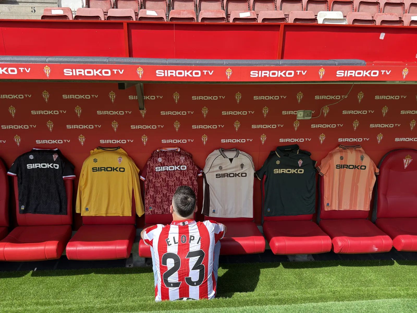 Elopi23 with back to camera wearing ELOPI 23 shirt on the El Molinón bench, with Sporting 2026/27 kits hung up