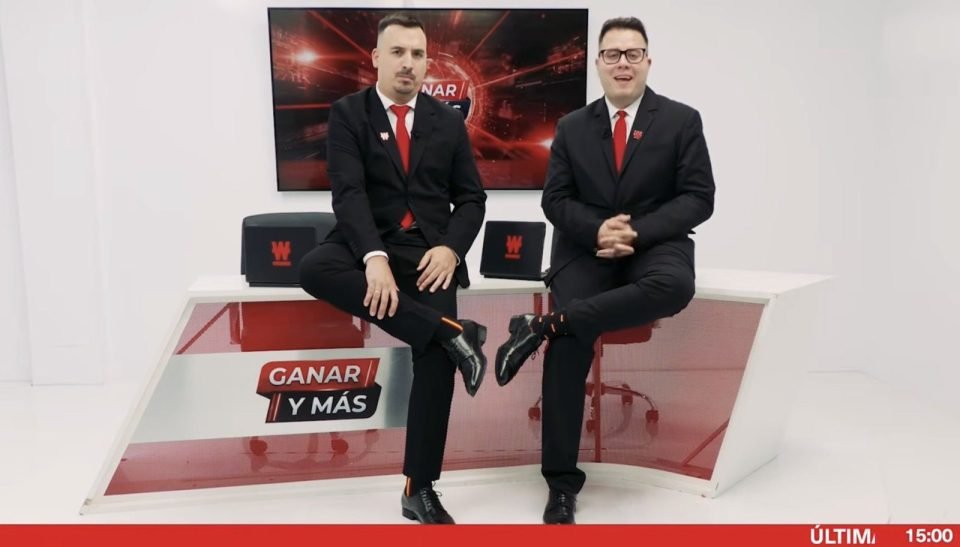 Los dos presentadores de Ganar y Más, de traje negro y corbata roja, sentados sobre la mesa del plató con el rótulo Ganar y Más